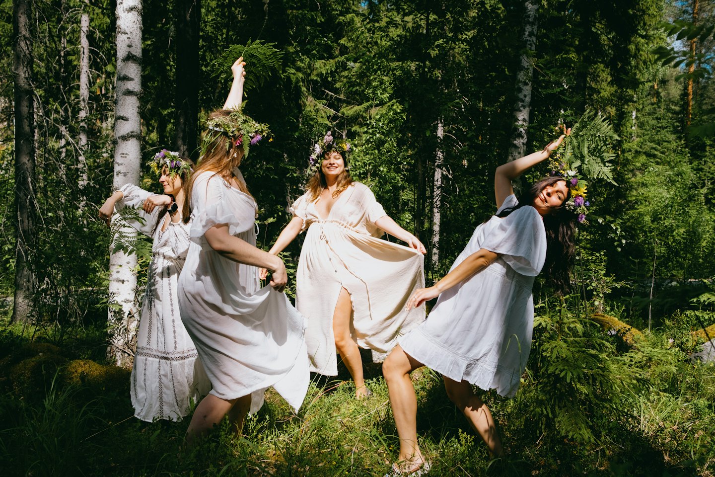 Quatre femmes couronnées de fleurs dansent ensemble dans une forêt.
