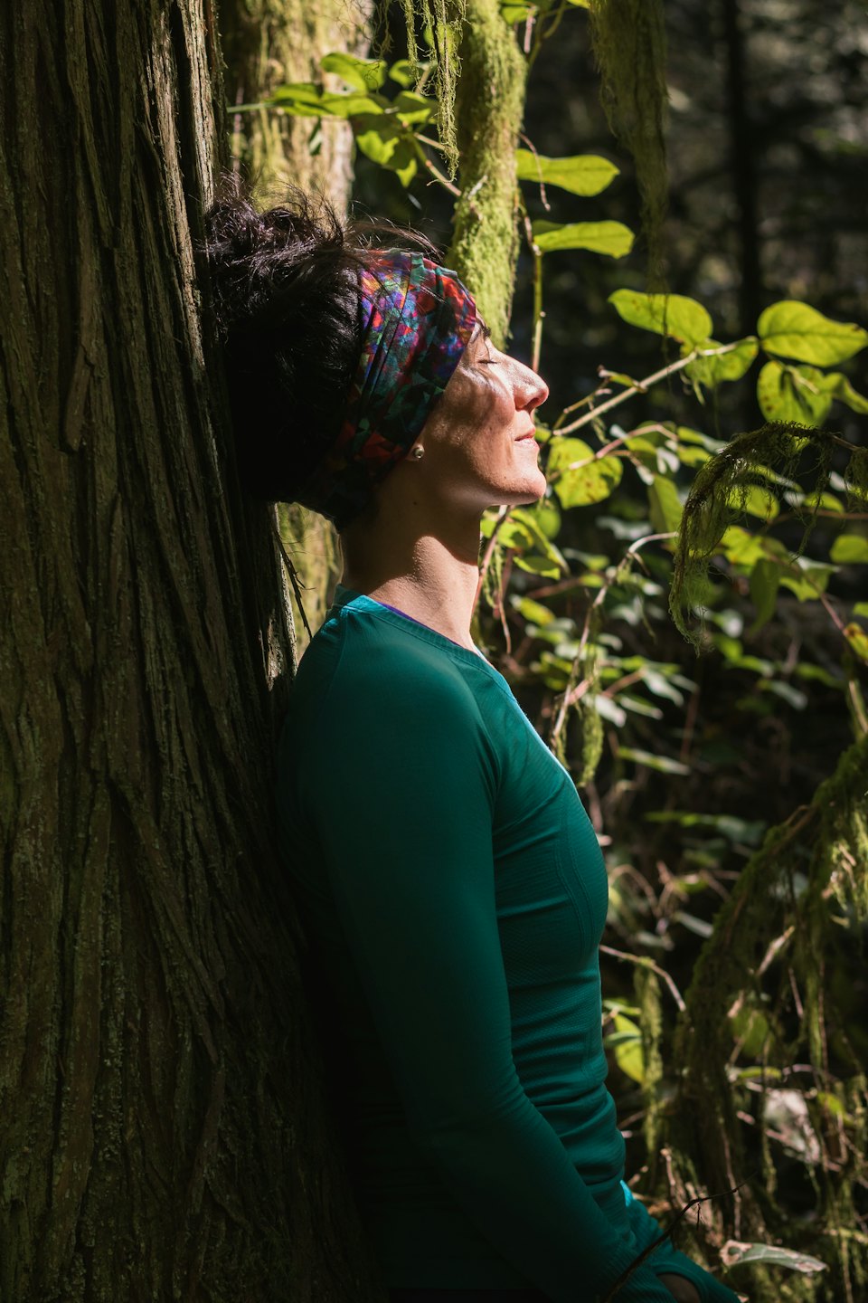 Une femme, les yeux fermés, s'appuie contre un arbre couvert de mousse dans une forêt.